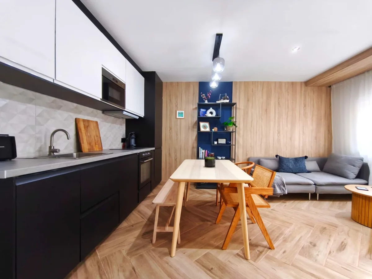 Vista general del apartamento: cocina negra al fondo, mesa de comedor de madera clara en el centro y el sofá a la derecha, todo en la misma pieza y con el mismo suelo de espiga.